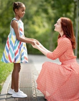 Woman wearing a daytime dress, kneeling and holding the hands of a pre-teen.