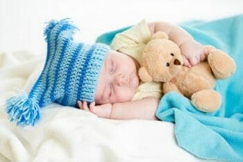 Adorable baby sleeping with a blue, knitted hat holding a Teddy bear.