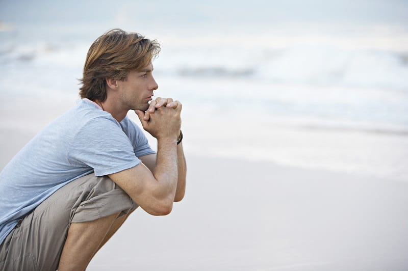 Young man crouching by the sea shore looking at sea, thoughtful.
